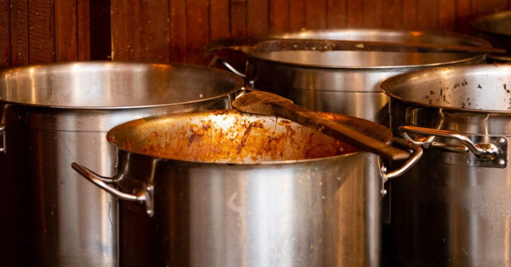 Stainless steel cooking pots with wooden spatula on a stove in a restaurant kitchen.